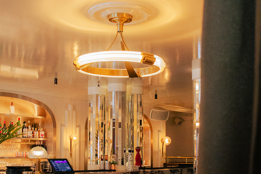 Suspension circulaire dorée rétro-éclairée au-dessus d’un comptoir de bar chic, entourée de colonnes en miroirs et d’un décor intérieur art déco lumineux