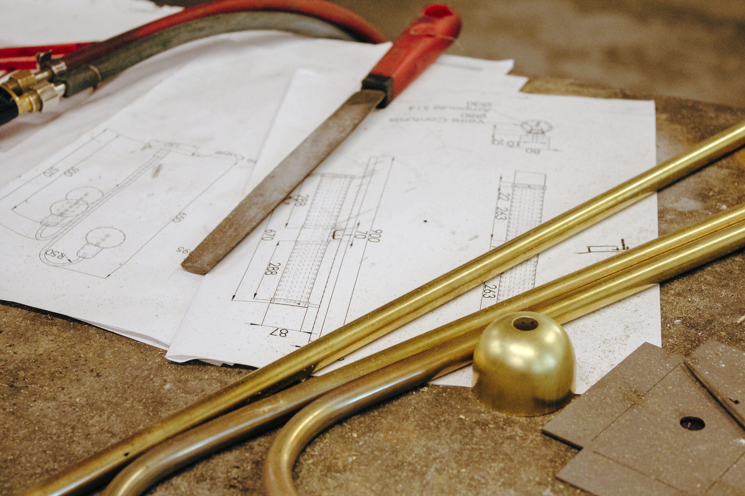 Plan technique de luminaire avec pièces en laiton, lime et chalumeau posés sur un établi d’atelier, illustrant la phase de conception et de fabrication artisanale en design industriel