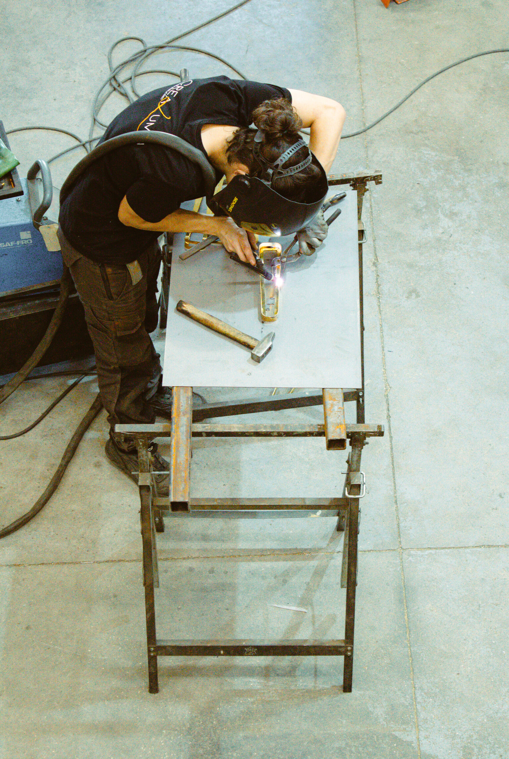Soudeur en plein travail dans un atelier industriel, utilisant un poste à souder TIG sur une pièce métallique fixée sur un établi, illustrant un savoir-faire artisanal de précision