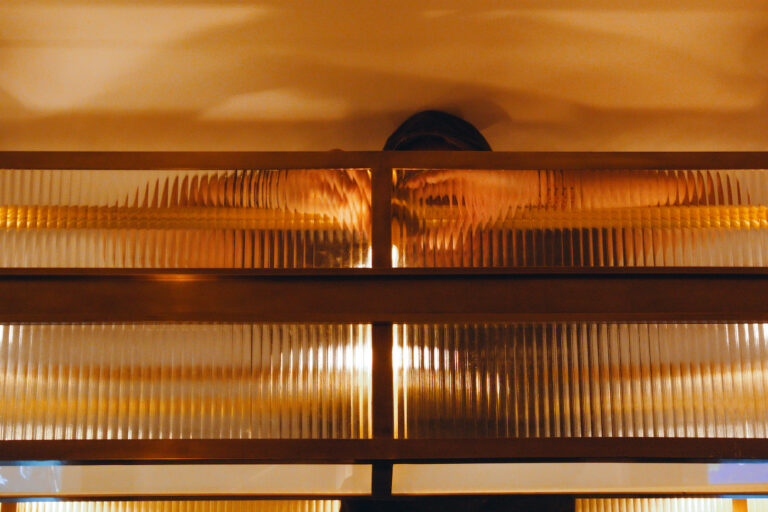 Pose d’un luminaire derrière une verrière en verre prismatique et structure en laiton, reflétant la lumière et les mains du monteur à travers les lignes striées du verre