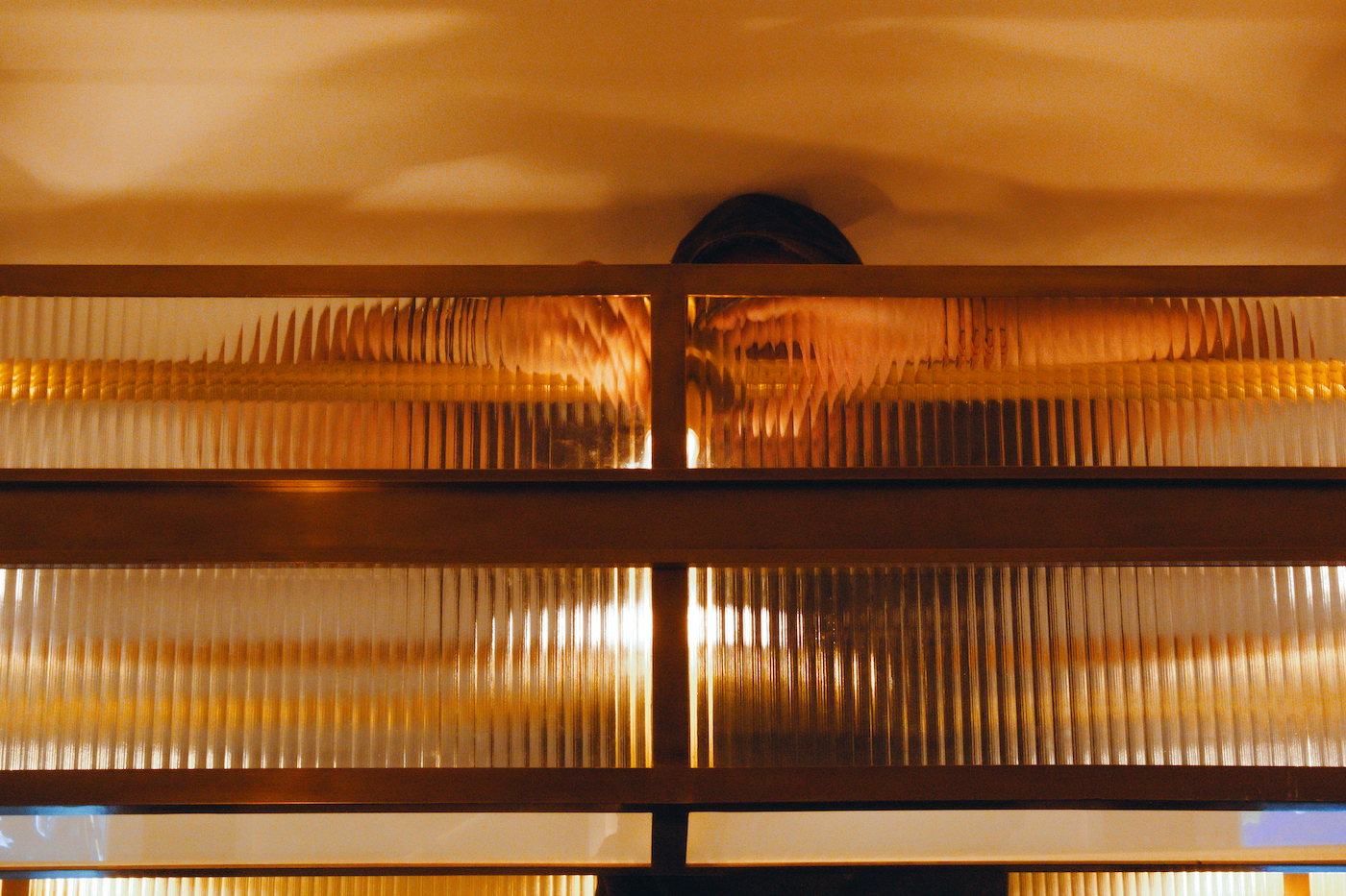 Pose d’un luminaire derrière une verrière en verre prismatique et structure en laiton, reflétant la lumière et les mains du monteur à travers les lignes striées du verre