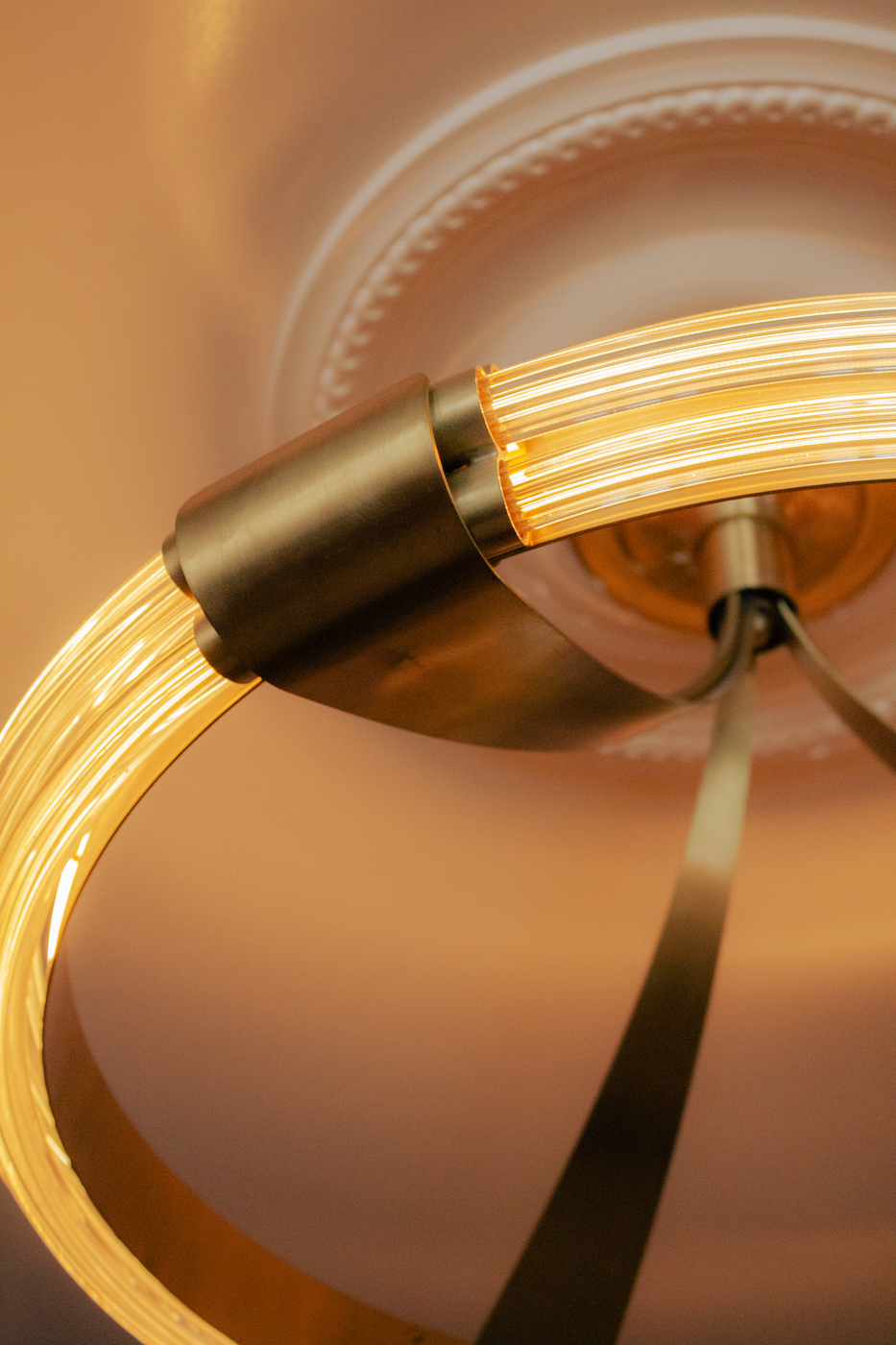 Gros plan sur un luminaire circulaire en laiton et tubes lumineux incurvés, suspendu sous une rosace de plafond en plâtre décoratif