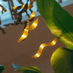 Suspension lumineuse en rubans de laiton ondulés suspendus au plafond, évoquant des éclats de lumière flottant parmi des feuilles tropicales en intérieur sous verrière