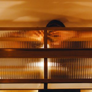 Pose d’un luminaire derrière une verrière en verre prismatique et structure en laiton, reflétant la lumière et les mains du monteur à travers les lignes striées du verre